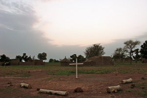 A simple cross sits in the ground with buildings in the background.