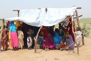 A group of people are gathered closely together under a rudimentary shelter made of poles and white cloth.