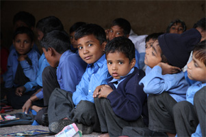 A group of schoolboys is sitting on the floor. Some are looking at the camera, others are looking at the teacher.