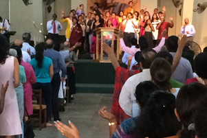 A congregation and a group at the front are worshipping the Lord together, many with their hands raised. 