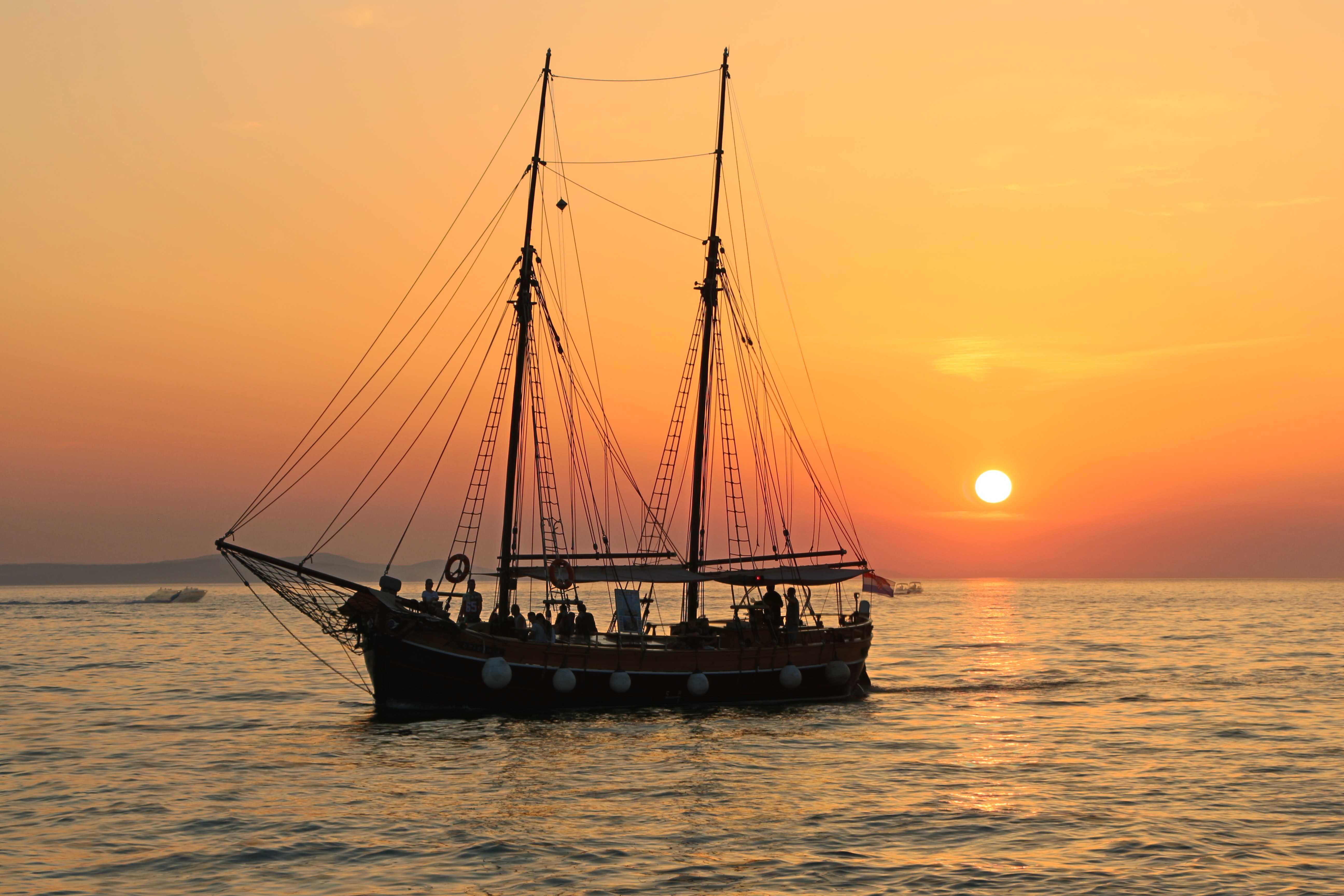 A scene of an antique ship sailing across the water, a bright orange sunset on the horizon in the background.