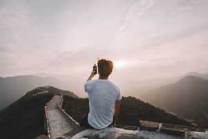 A young man sitting on the Great Wall of China, taking a picture with his cell phone.