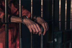 Man's hands extending from prison bars with handcuffs on his wrists.