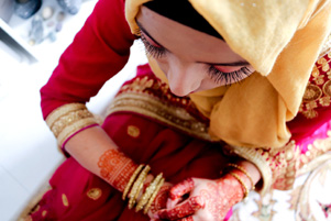 A Muslim bride sits with her head down. She is dressed in brightly coloured clothing and henna decorations cover her hands.