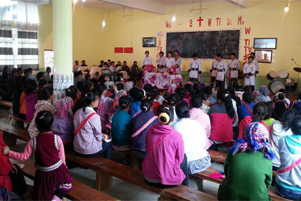 An audience is seated on wooden benches facing some individuals at the front of the meeting.