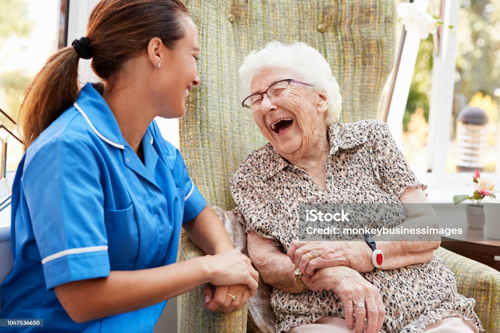 senior-woman-sitting-in-chair-and-laughing-with-nurse-in-retirement-picture-id1047536650